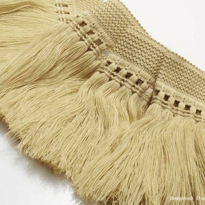 May include: A beige decorative fringe trim featuring a wide, textured woven header with a repeating geometric pattern. Attached to the header is a row of long, straight, silky tassels that hang vertically, creating a uniform and dense appearance.