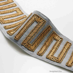 May include: Gray fabric trim with gold beaded rectangular shapes. The trim is beaded with small gold beads in a chevron pattern.
