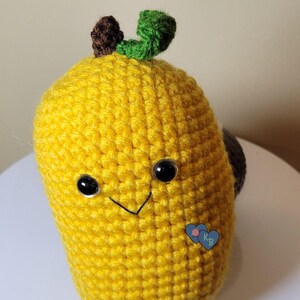Puede incluir: Un mango amarillo de ganchillo hecho a mano con ojos de seguridad negros y una sonrisa cosida. El mango tiene un tallo marrón y una hoja verde. Una pequeña etiqueta azul en forma de corazón con las letras "KB" está adjunta al costado.