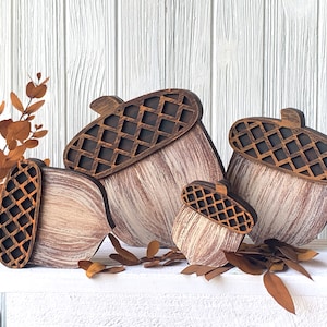 May include: Four wooden acorns of varying sizes, painted in a whitewashed style with brown accents. The acorns are arranged in a staggered formation, with the largest acorn at the back and the smallest acorn in the front. The acorns are a popular fall decor item.