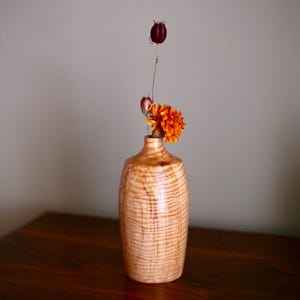 May include: A wooden vase with a light brown, textured finish, holding an orange flower and a dried seed pod. The vase sits on a dark wooden surface, with a neutral gray background.