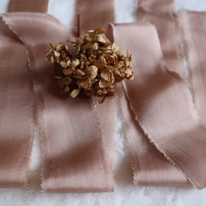 Peut inclure: Un ensemble de ruban de soie rose poussiéreux aux bords effilochés. Le ruban est disposé en éventail avec un petit bouquet de fleurs séchées au centre.