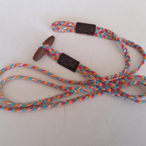 May include: A multi-coloured braided lanyard with a wooden toggle and leather details. The lanyard has a vibrant pattern of pink, orange, blue, and white threads. The leather details are brown.
