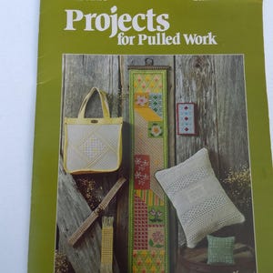 May include: A green leaflet titled "Projects for Pulled Work" featuring various needlework projects. The projects include a tote bag, a long wall hanging, a small pillow, and a decorative belt. The background is a rustic wooden wall.