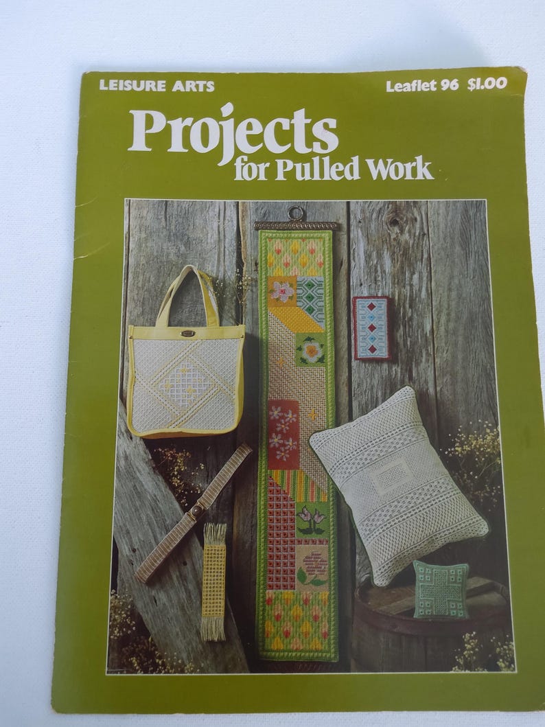 May include: A green leaflet titled "Projects for Pulled Work" showcasing various needlework projects. The projects include a tote bag, a long wall hanging, a small cushion, and a decorative belt. The background is a rustic wooden wall.