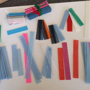 May include: A collection of plastic canvas strips and sheets in various colors, including blue, pink, green, red, and white. Some strips are bundled together with twine. These craft supplies are ideal for needlepoint and other DIY projects.