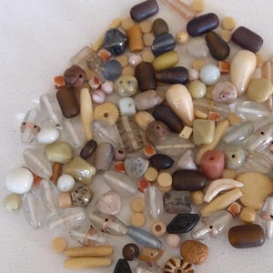 May include: A collection of assorted beads in various shapes, sizes, and colors. The beads include clear, brown, beige, and pastel-colored options. Some beads are cylindrical, while others are rounded or faceted. The beads are scattered on a white surface.