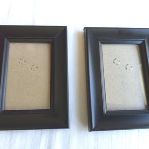 May include: Two rectangular picture frames with a smooth matte black finish. Each frame features a beveled inner edge leading to a clear glass pane, which protects a light tan cardboard backing insert. The frames are oriented vertically.