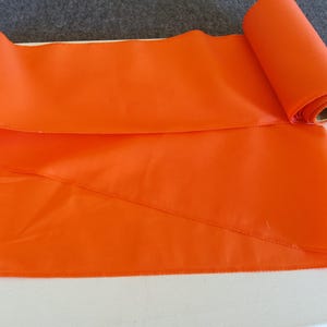 May include: A vibrant orange fabric roll, partly unrolled, showing a smooth, satin-like texture. The material is placed on a white surface, with a section rolled up, exposing the cardboard core. Suitable for crafting or decorative purposes.