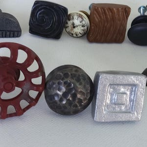 May include: A selection of decorative cabinet knobs in various styles and finishes, including a red metal knob with a circular design, a silver metal knob with a square design, a black metal knob with a hammered finish, a grey metal knob with a half-circle design, a brown ceramic knob with a woodgrain pattern, a black ceramic knob with a round design, a white ceramic knob with a clock face design, and a grey ceramic knob with a swirl design.