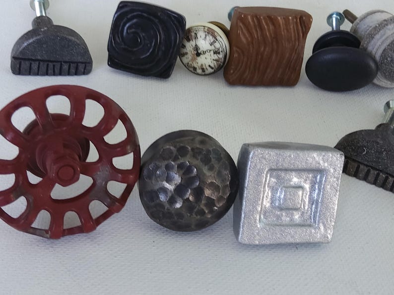 May include: A selection of decorative cabinet knobs in various styles and finishes, including a red metal knob with a circular design, a silver metal knob with a square design, a black metal knob with a hammered finish, a grey metal knob with a half-circle design, a brown ceramic knob with a woodgrain pattern, a black ceramic knob with a round design, a white ceramic knob with a clock face design, and a grey ceramic knob with a swirl design.