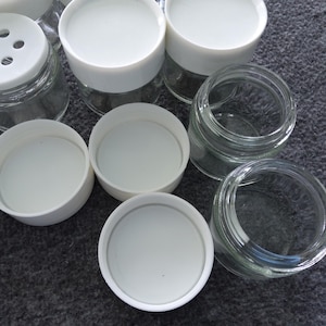 May include: A collection of small, clear glass jars with white plastic lids. Some lids are on the jars, while others are separate. One lid has holes for dispensing. The jars are on a gray surface.