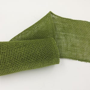 May include: A roll of olive green burlap ribbon featuring a loose, open-weave mesh texture. The fabric is partially unrolled, displaying a consistent grid pattern throughout the material with frayed, unfinished edges on the sides of the strip.