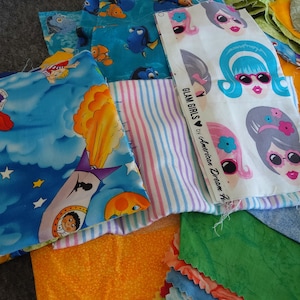 May include: A collection of colorful fabric scraps. Prints include cartoon characters, striped patterns, and faces with sunglasses. The fabric pieces are in shades of blue, orange, green, and pink.