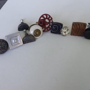 May include: Assortment of decorative cabinet knobs in various styles and colours, including a hammered metal knob, a square knob, a ceramic knob with a floral design, a metal knob with a clock face, and a wooden knob with a wood grain pattern.