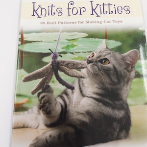 May include: A softcover book titled Knits for Kitties: 25 Knit Patterns for Making Cat Toys by Sara Elizabeth Kellner. The cover features a grey tabby cat reaching for a knitted dragonfly toy suspended by a purple yarn string.