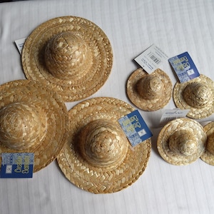 May include: A collection of six round, woven straw hats in varying sizes, each featuring a raised crown and a flat brim. Several of the hats have blue and white rectangular product tags attached, with some tags displaying barcodes and text.