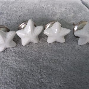 Op de afbeelding: Vier witte keramische zeesterkastknoppen met zilveren beslag. De knoppen zijn in een rij gerangschikt, elk met een vijfpuntsontwerp. Op een grijze, getextureerde ondergrond.