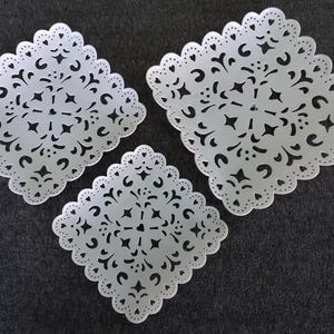 May include: Three white, square-shaped decorative trays with intricate, openwork floral and geometric patterns. The trays have scalloped edges and are arranged on a dark gray surface, creating a visually appealing display.