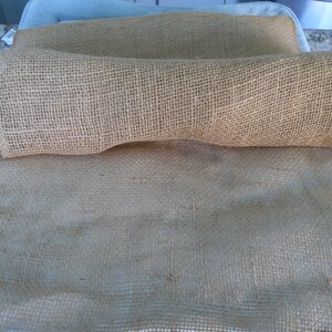 Könnte beinhalten: Nahaufnahme von gerolltem und entfaltetem Jute-Stoff. Der naturfarbene Jute ist grob und gewebt, mit einem sichtbaren Gittermuster. Der Stoff liegt auf einer weißen Oberfläche, was auf einen Bastel- oder Dekorationsartikel hindeutet.