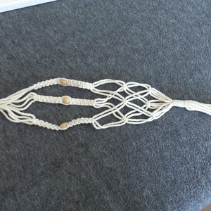 May include: Off-white macrame plant hanger with a wooden ring and three small wooden beads. The knotted design features a diamond-shaped pattern in the center and a tassel at the bottom. The plant hanger is designed to hold a small to medium-sized plant pot.