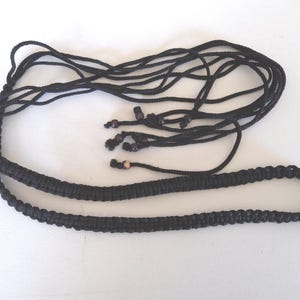 May include: A black macrame belt with a braided design. The belt features a long, braided section and several loose cords with decorative beads at the ends. The belt is displayed on a white surface.