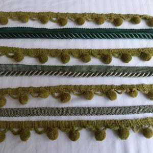 May include: A collection of seven horizontal strips of decorative trim arranged in parallel. The set includes four olive green pom-pom fringes, one dark green twisted cord trim, one sage green braided trim, and one metallic silver gimp trim.