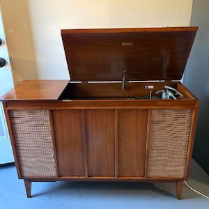 May include: A wooden record player console featuring a hinged top lid that opens to reveal a turntable mechanism. The front of the cabinet displays two side panels with woven cane speaker grilles and a central section with vertical wood slats.