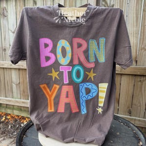 May include: A dark grey t-shirt with the slogan "BORN TO YAP!" in colourful, playful lettering. The shirt features the brand name "Heather Nicole Company" at the top. The design includes stars and a variety of bright colours.