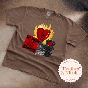May include: A brown t-shirt featuring a graphic design with a red heart, flames, a record, and a traffic light. The text "RED BEDROOM RECORDS" is on a red rectangle. The shirt also has the text "Heather Nicole Company".
