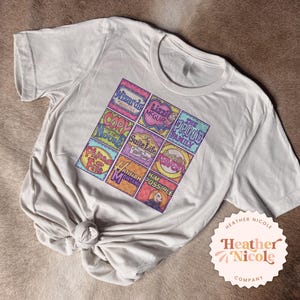 Könnte beinhalten: Weißes T-Shirt mit einer bunten Grafik von Logos beliebter Disney Channel-Shows, darunter 'Lizzie McGuire', 'Hotel Zack & Cody', 'Hannah Montana', 'Kim Possible', 'Die Proud Family', 'Cory in the House', 'Die Zauberer vom Waverly Place' und 'Phineas und Ferb'.