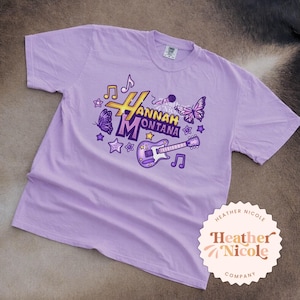 May include: A light purple t-shirt featuring a graphic design with the text "Hannah Montana" in gold and purple. The design includes musical notes, stars, a guitar, and butterflies. A circular logo with the text "Heather Nicole Company" is in the bottom right corner.