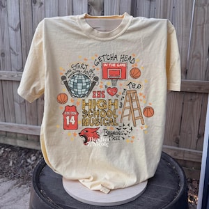 May include: A light yellow t-shirt with a graphic design featuring a basketball hoop, basketballs, and the text "EHS High School Musical." The design also includes a jersey with the number 14 and the word "Bolton."