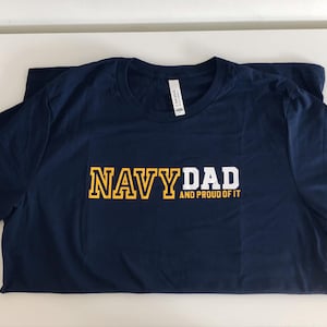 May include: Navy blue t-shirt with the text "NAVY DAD AND PROUD OF IT" in yellow and white lettering.