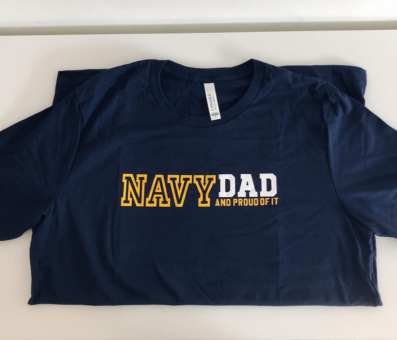 May include: Navy blue t-shirt with the text "NAVY DAD AND PROUD OF IT" in yellow and white lettering.