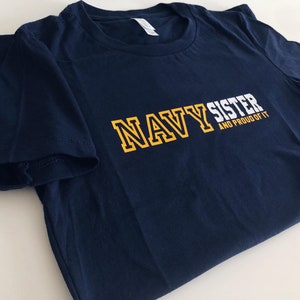 May include: Navy blue t-shirt with the text "NAVY SISTER AND PROUD OF IT" in white and yellow lettering.