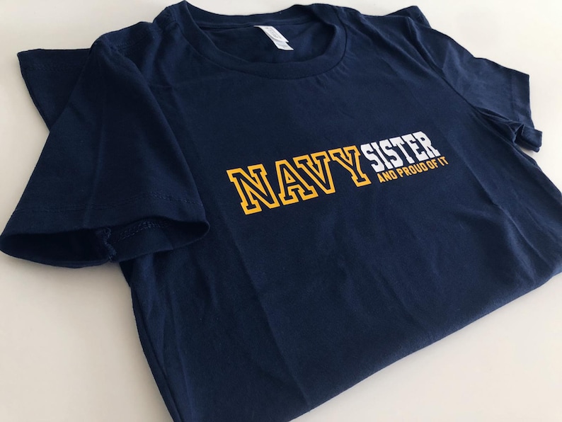 May include: Navy blue t-shirt with the text "NAVY SISTER AND PROUD OF IT" in white and yellow lettering.