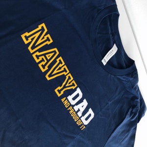 May include: Navy blue t-shirt with the text "NAVY DAD AND PROUD OF IT" in white and yellow lettering.