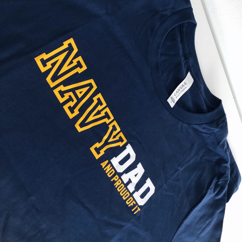 May include: Navy blue t-shirt with the text "NAVY DAD AND PROUD OF IT" in white and yellow lettering.