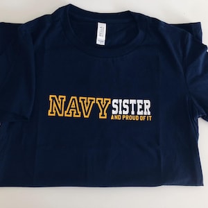 May include: Navy blue t-shirt with white text that reads "Navy Sister and Proud of It". The word "Navy" is in yellow outlined letters.