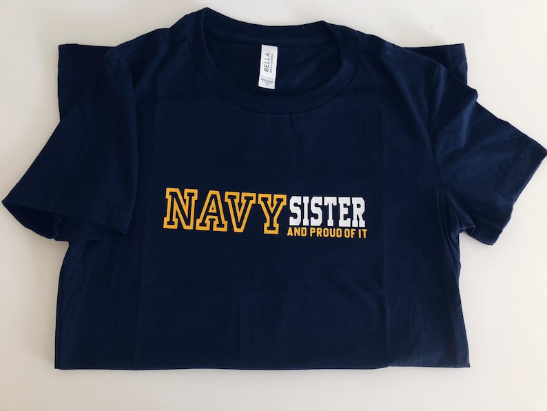 May include: Navy blue t-shirt with white text that reads "Navy Sister and Proud of It". The word "Navy" is in yellow outlined letters.