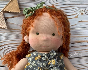 May include: A handmade doll with red curly hair wearing a gray floral dress with a green bow. The doll is sitting on a white wooden background with a banner of fabric triangles.