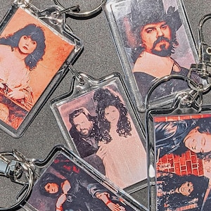 May include: Five different keychains with colorful images printed on them. The keychains feature a woman with long dark hair, a man with a beard and a hat, a man and a woman, and two women with dark hair.