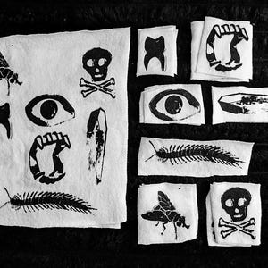 Könnte beinhalten: Schwarz-Weiß-Illustrationen verschiedener gotischer und makabrer Symbole. Die Bilder zeigen einen Totenkopf mit gekreuzten Knochen, eine Fliege, ein Auge, einen Zahn, einen Tausendfüßler, einen Sarg und Reißzähne. Die Illustrationen sind auf kleinen, quadratischen Papierstücken gedruckt.
