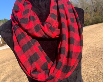 Könnte beinhalten: Ein rot-schwarz karierter Loop-Schal, präsentiert auf einer schwarzen Schaufensterpuppe. Der Schal besteht aus einem weichen Stoff und ist kreisförmig drapiert. Der Hintergrund zeigt eine sonnige Szene im Freien.