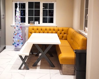 May include: A white table with a black X-shaped base sits in a dining nook with a yellow tufted banquette. A small white Christmas tree with colorful ornaments is in the background. A neon sign with the word "Slainte" is on the wall.