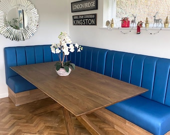 May include: A blue, upholstered dining bench with a wooden base and a wooden table with a cross-shaped base. The table is set in front of the bench. The wall behind the bench has a sign that reads "LONDON BRIDGE" and "KINGSTON".