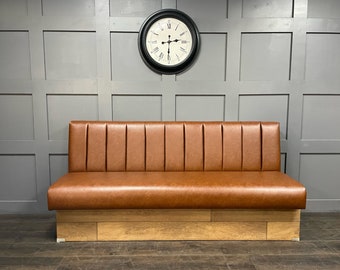 May include: A brown leather bench with a wooden base against a gray wall with a black clock. The bench has a tufted back and a curved front.
