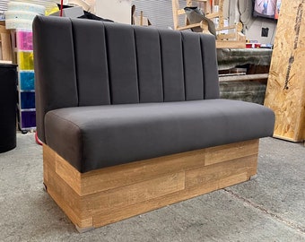 May include: A gray upholstered booth with a wooden base. The backrest is vertically channeled. The base is constructed of light brown wooden planks. The booth is designed for seating in a restaurant or cafe setting.