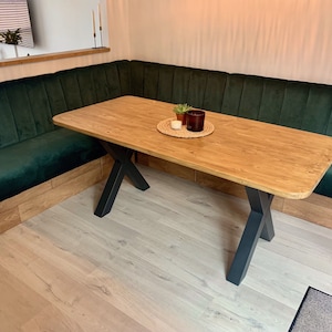 May include: A green velvet corner booth with a wooden table and black X-shaped legs. The table has a light wood finish and is set with a small potted plant and a candle.